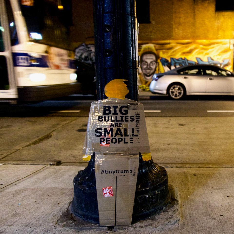 tiny trump on a sidewalk at night with the slogan 'Big Bullies Are Small People'